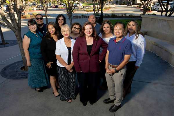 Sandra Armenta stands with a group of community members outdoors for a group portrait in a plaza, with trees, walkways, and campus buildings in the background.