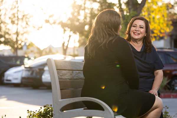 Sandra Armenta smiles while speaking with another person seated across from her on an outdoor bench, with warm evening sunlight filtering through trees in the background.