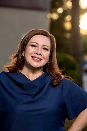Portrait of Sandra Armenta smiling at the camera while wearing a burgundy blazer over a black top, standing outdoors with a softly blurred background of green trees.